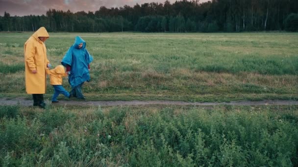 Wieczorem rodzina z dzieckiem spaceruje po parku. Ludzie noszą płaszcze przeciwdeszczowe. — Wideo stockowe