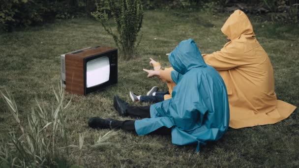 Rodina sedí na trávě v pláštěnkách a dívá se na starou retro televizi — Stock video