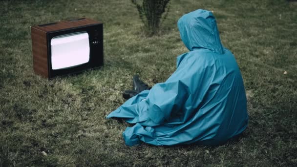 フード付きブルーレインコートの女性は草の上に座っていると古いレトロテレビを見て — ストック動画