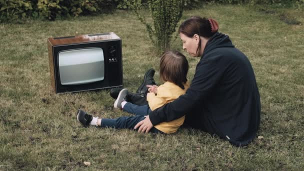 Žena, matka s malým dítětem sedí na trávě a dívají se na starý retro TV — Stock video