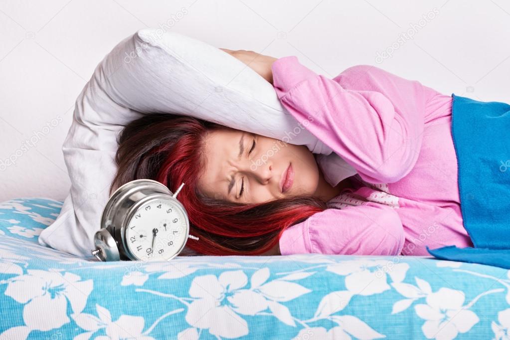 Annoyed girl covering her ears with a pillow from the alarm clock