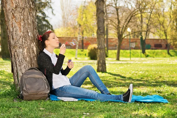 Ung flicka lutar mot ett träd blåser tvål bubblor — Stockfoto