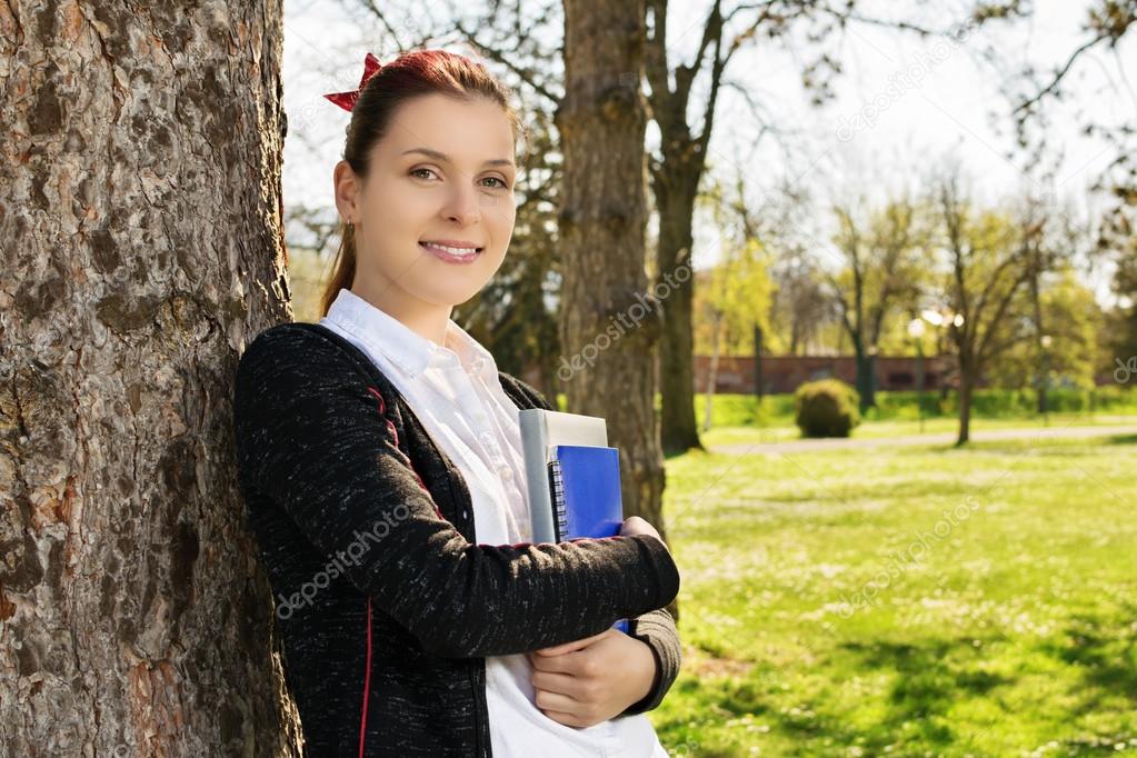 Female student leaning against a tree holding books