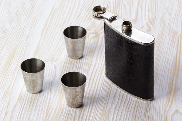Metal flask trimmed leather and three metallic, sturdy shot glasses. — Stock Photo, Image