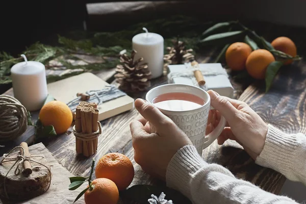 Woman drinks hot tea while preparing gift packages on wooden table. Chic cozy craft gift boxes. Natural aestetic. Zero waste, plastic free, trendy hand made gift package for Christmas.