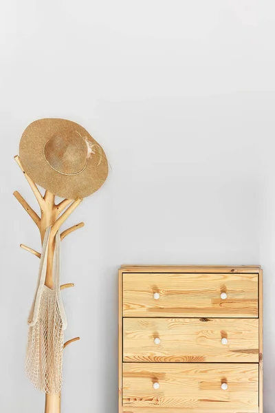 Minimalistic hallway interior with trendy wooden clothes hanger stand and commode against a gray wall.Wicker hat and mesh bag on the hanger.Copy space