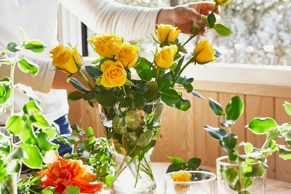 Женщина расставляет свежие весенние желтые цветы в стеклянной вазе на деревянном столе у окна. Желтые розы. Естественное освещение. Мягкий фокус. — стоковое фото