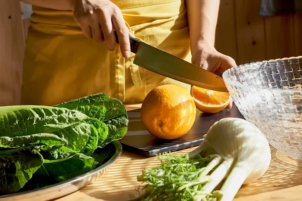 Kadın kesme tahtasında portakal kesiyor ve marul, rezene ve portakal salatası hazırlıyor. Vitamin ve sağlıklı beslenme. Doğal ışık. Seçici odak.