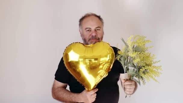 Handsom mature bearded man smiling and giving mimosa flower and heart ballon to viewer. Man with bouquet.Holiday concept — Stock Video
