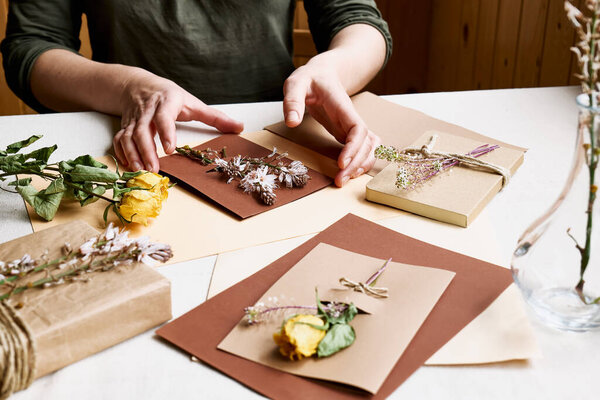 Woman makes zero waste, plastic free, trendy hand made gift card with craft recycled paper and dried flowers on the table with linen tablecloth. Natural aestetic.
