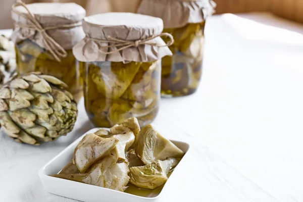 Artichokes hearts in glass jar with olive oil and herbs. Pickled artichoke with garlic in a glass jar on white tablecloth table. Homemade healthy eating