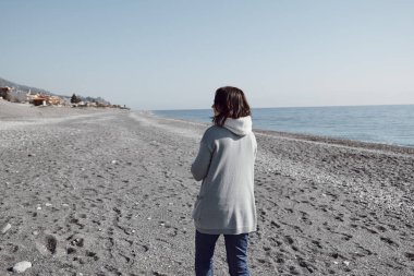 Terkedilmiş kış plajında yürüyen, güneşli havanın tadını çıkaran bir kadın.