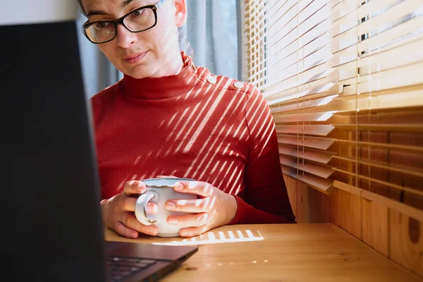 Woman wearing red turtleneck works with laptop near the window with shutters. Remote working concept. Selective focus