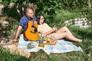 Orta yaşlı bir çift bahçede taze egzotik meyve ve tatlı sandviçlerle piknik yapıyor. Çiçek desenli mavi tişörtlü adam gitar çalıyor. Bahar, yaz zamanı yaşam tarzı, aşk randevuları..