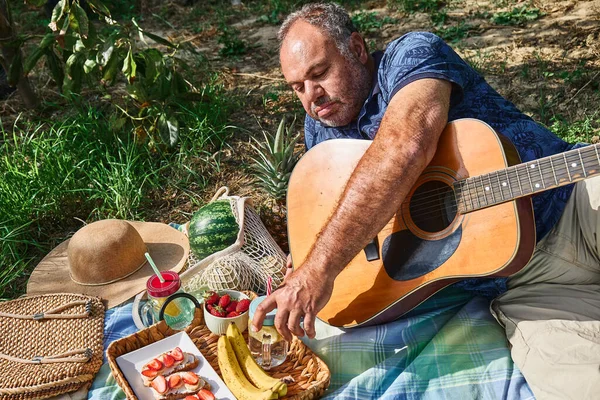 Middle aged couple having a picnic in the garden with fresh exotic fruit and sweet sandwiches. Man in blue t-shirt with flower pattern plays the guitar. Spring, summer time lifestyle, love dating