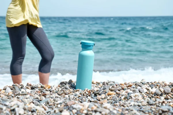 Güneşli bir yaz gününde çakıl taşı plajında, arkasında mavi deniz olan bir çelik termal şişe. Sağlıklı yaşam tarzı, su dengesi kontrolü, cilt bakımı konsepti. Seçici odak.
