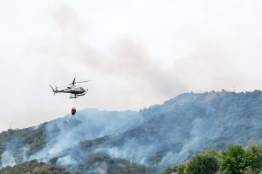Yangın helikopteri dağlardaki bir orman yangınında kovadan su bırakıyor. Orman yangını çevresel bir felakettir. Ormanların tahrip edilmesi.