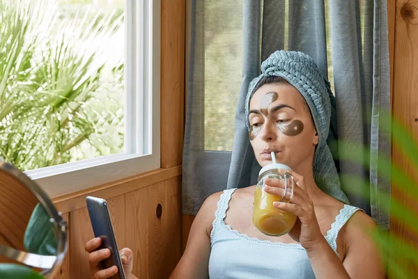 Skincare routine. Middle age woman with wrinkles and hydrogel under-eye patches drinking smoothie and looking in window. Lifting anti-wrinkle mask under eyes. Skincare, Collagen mask and spa concept.