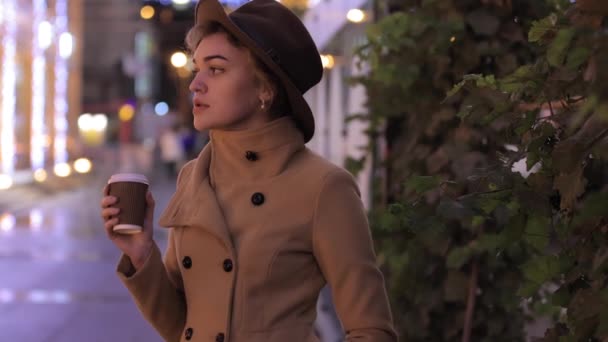 Piękna kobieta w kapeluszu i płaszczu pije kawę w nocnym mieście — Wideo stockowe