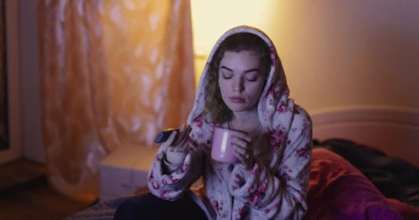 Žena sedí v posteli a dívá se na televizi a pije čaj — Stock video