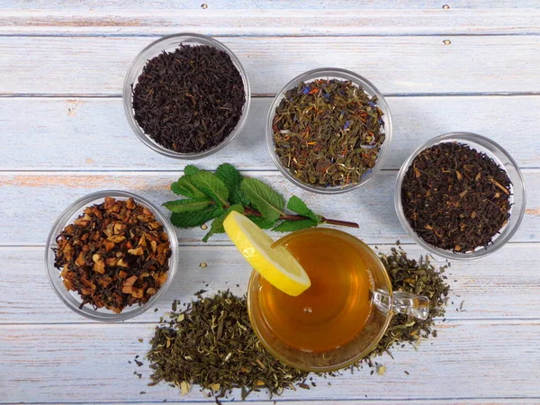 Green tea cup with a lemon wedge and mint leaves, accompanied by bulk tea and various types of tea in bowl, on a vintage wood background