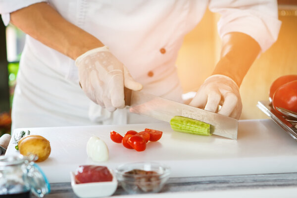 Hand with knife cutting cucumber. White cooking board with vegetables. Lets cook a salad. Only high quality ingredients.