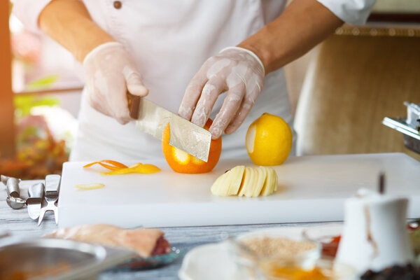 Hand with knife peels orange. Orange on cooking board. Chef needs some orange rind. Ingredient for meat dish.