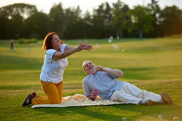 Happy mature couple blowing bubbles lying on the field. — Stock Photo, Image