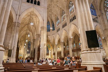 New York City, New York, ABD - 07.31.2024: Bir grup ziyaretçi yüksek kemerli ve boyalı camlı pencereli büyük bir Gotik katedralin içini keşfediyorlar. Karmaşık taş sütunlar ve detaylı mimari görkemli ve kutsal bir yapı yaratır.