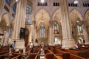 New York City, New York, ABD - 07.31.2024: Ziyaretçiler yüksek kubbeli tavanları ve boyalı pencereleri olan çarpıcı bir Gotik katedralin içinde yürüyorlar. Mimari taş kemerler, sütunlar ve dini dekorlarla görkemli..