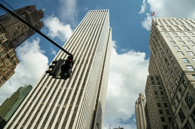 Manhattan gökdelenlerinin yukarıdaki manzarası. Parlak mavi bir gökyüzü ve dağınık bulutlar altında. Ön planda bir kırmızı trafik ışığı askıya alınarak, yüksek binalara kontrastlık eklendi.