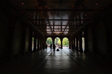 New York City, New York, ABD - 07.31.2024: İnsanlar gölgeli Bethesda Terrace Arcade 'den Central Park' taki parlak çeşme plazasına doğru yürüyorlar. Kemerli tavan ve fayanslı zemin bu tarihi New York 'ta dramatik bir siluet sahnesi yaratıyor.