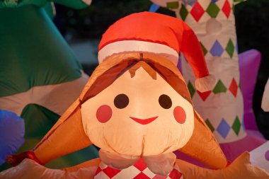 Gülümseyen, şişme bir kız Noel Baba şapkası takıyor ve kırmızı-beyaz bir kıyafet giyiyor. Sıcak tatil ışıkları gece sevimli karakterin etrafında parlıyor..