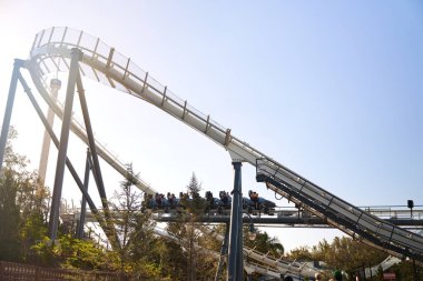 Dik bir düşüş ve parlak arka plan boyunca kesilen uzun kolonlar. Eğimli raylar, ağaçlar ve gökyüzü dramatik bir tema parkı sahnesi.