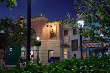 Orlando, Florida, ABD - 11.15.2024: Bir tema parkındaki tuğla ve sıva binalar Noel çelenkleri ve alacakaranlıkta parlayan sokak lambalarıyla süslenmiştir. Yeşil ağaçlar ve çalılar sıcacık şehir tatil sahnesini çerçeveliyor.