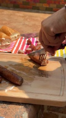 Ahşap bir kesme tahtasının üzerinde ızgara sosisi bıçak ve çatalla doğrayan bir elin yakın çekimi. Ekmek ve renkli tabaklar açık hava barbekü atıştırmalığı sırasında yakınlarda otururlar..