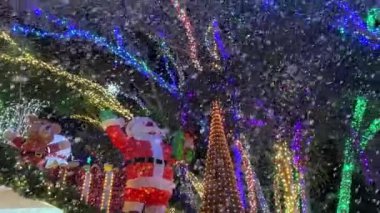 Parlayan ağaçların altında oyuncak ayı süslemesinin yanında hediye tutarken şişme Noel Baba dalgaları. Renkli ışıklar ve yüzen baloncuklar gece açık havada şenlikli bir kış manzarası yaratır..