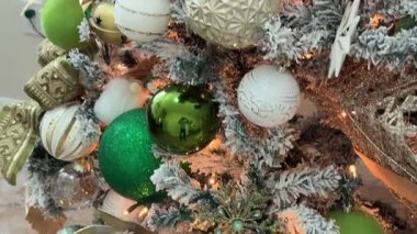 Yeşil ve beyaz takılar, kurdele ve sıcak peri ışıklarıyla süslenmiş bir Noel ağacının yakınında. Parlak süsler kapalı alanda neşeli bir tatil ortamı yaratır..