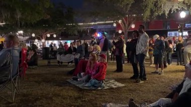 Winter Park, Florida, ABD - 12.14.2024: Karmaşık bir kalabalık gece bir kasaba meydanında canlı performans izlemek için çimlerin üzerinde toplanıyor. Tatil ışıkları ve mağazalar sıcak bir topluluk ortamı yaratıyor..