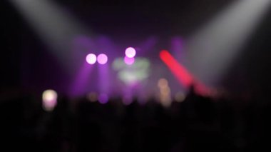 Mor spot ışıkları ve güçlü bir kırmızı ışıkla bulanık bir konser sahnesi. Kalabalık siluetler ve parlayan bir telefon ekranı hayranları performansı kaydediyor.