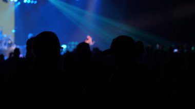 Siluetli konsere gidenler, üzerinde parlak ışıklar bulunan mavi ışıklı bir sahnede bir sanatçıyı izliyorlar. Bazı insanlar canlı müzik şovunu kaydetmek için telefonları soyuyor..