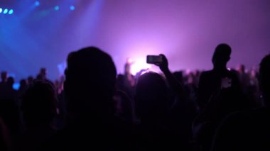 Siluetli seyirciler mor ve mavi sahne ışıkları altında bir konser izliyorlar. Kalabalık mekanda bir kişi video kaydetmek için akıllı telefonu soyuyor..