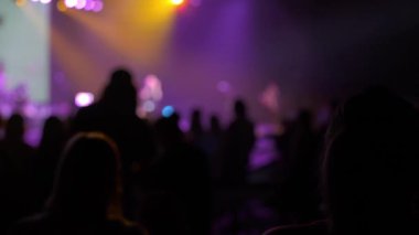 Kalabalığın odaklanmamış siluetleri mor ve sıcak sarı spot ışıklarıyla aydınlatılmış bir sahneye bakıyor. Bulanık bokeh ve duman canlı bir müzik olayının enerjisini yakalıyor.