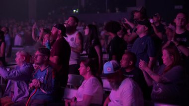 Orlando, Florida, ABD - 08.17.2025: Bankta ve koridorlarda loş mor ışık altında kalabalık bir konser salonu. Seyirciler sahnede canlı performansı beklerken ayağa kalkar, oturur ve telefon kullanır..