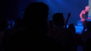 Büyük bir siluet ve kaldırılan eller, mavi ışıklı bir sahnede şarkı söyleyen bir müzisyeni çerçeveletir. Kalabalık canlı müzik etkinliği sırasında dumanlı bir mekanda tezahürat yapıyor..