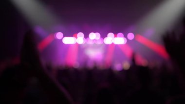 Mor sahne ışıkları ve kırmızı spot ışıklarıyla soyut bulanık konser sahnesi. Büyük bir seyirci sahnenin önünde siyah siluetler olarak belirir..