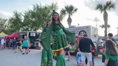 Orlando, Florida, ABD - 03.16.2024: Yeşil kostümlü gülümseyen bir sanatçı bir Aziz Patrick Günü festivalinde kalabalığı eğlendiriyor. İnsanlar açık hava kutlamaları sırasında yemek kamyonları ve çadırların etrafında toplanıyor..