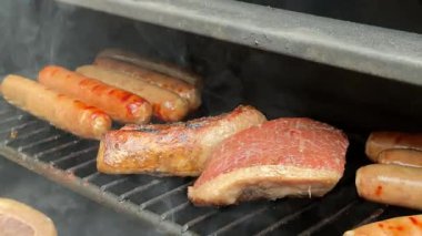 Mangal ızgarasının üst rafında dolgun sosisler ve dana picanha dumanı. Olay yerinde, evde yapılan piknik sırasında kahverengi kaplamalar, yağlar ve yükselen buhar görülüyor..