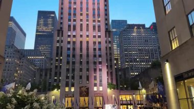 New York, New York Eyaleti, ABD - 07.29.2024: Manhattan şehir merkezindeki Rockefeller Center 'daki uzun ofis kuleleri mavi saatte parlıyor. Aydınlatılmış pencere ve bayraklar açık bir akşam gökyüzünün altındaki şehir meydanında..