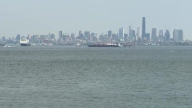 New York şehrinin ufuk çizgisi ufukta kargo gemileri ve limandaki teknelerin arkasında uzanıyor. Geniş su önplanı, işlek liman ve kentsel liman sınırlarını vurguluyor.